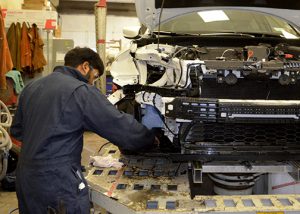 A student wearing coveralls works on the engine of a car.