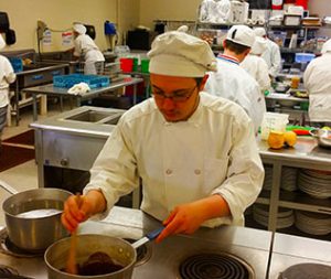 Male student in chef's hat and coat stirs a pot of something yummy in a kitchen setting. 