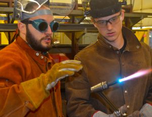 Teacher Don Mattoon wears goggles and headgear while instructing a welding student