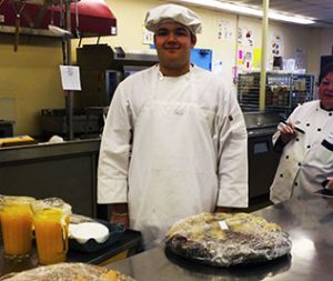 Student in chef's attire stands behind breakfast foods in a kitchen setting.