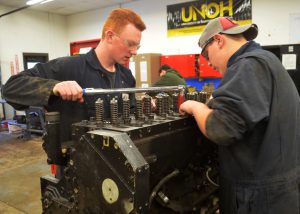 Two students dressed in coveralls work together on the engine of a diesel vehicle.