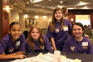 A group of girls in purple shirts smile as they speak about how they plan STEAM day events at their school.