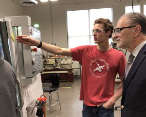 A young man in a red t-shirt and protective eyewear shows of a machine during a tour at the Center for Advanced Technology at Mohonasen.