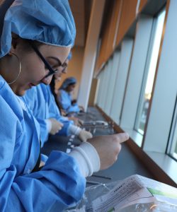 Students dressed in blue scrubs inspect medical equipment as part of a lesson on sterile processing.