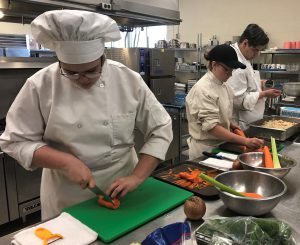  Career and Technical School culinary students Kathryn Manchester and Alexia Torres, dressed in chef’s uniforms, stand side by side and cut vegetables.