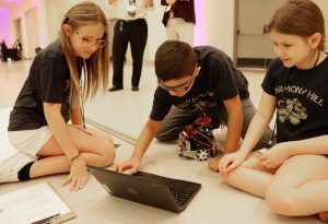 Three elementary age students work together at a laptop computer placed on a floor.