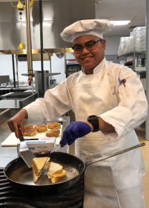Culinary student Angelo Carino, wearing a chef's uniform, smiles for the camera as he prepares a grilled cheese sandwich.