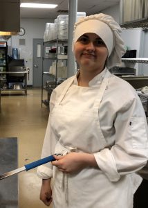 Culinary student Ginger Crandall, wearing a white chef's uniform, smiles for the camera while she cooks.