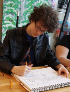 Capital Region BOCES graduate Tyler Blackman, wearing a black leather jacket and sitting before green and white backdrop, sketches scenery in a notebook.