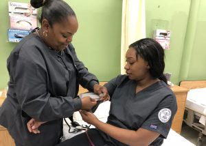 An Adult Nursing student practices taking a blood pressure with a classmate. Both women wear dark blue nursing scrubs.