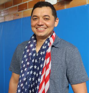 Adult Education student Javier Villatoro, wearing a blue short sleeved polo and an American flag scarf, smiles for the camera.