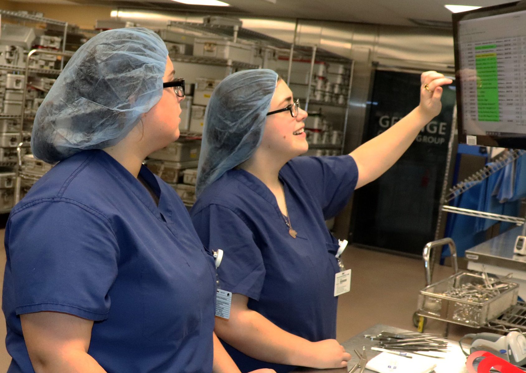 Two students in scrubs and hair coverings look into an overhead computer screen.
