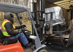 An adult student wearing a reflective vest unloads machinery using a forklist.