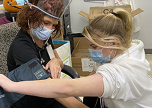A seated teacher, with curly red hair and wearing a protective blue face mask and clear face shield and black top has their blood pressure taken by a nursing student, who has blond hair, worn in a ponytail, and is wearing a blue face mask and white hoodie.