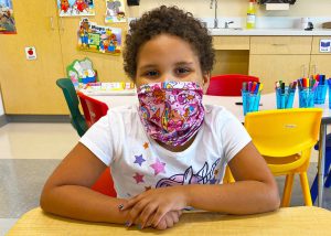 A young student wearing a pink printed protective face mask and sitting at a desk and smiles for the camera.