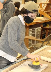 Student Cora Tribunella, wearing a tan cap, protective eyewear and a face mask, uses an orbital sander to to smooth a piece of wood in a Capital Region BOCES Building Trades classroom.