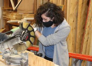 Student Isabel Tribunella, wearing protective eyewear and a face mask, uses a miter saw to cut through a board in a Capital Region BOCES Building Trades classroom.
