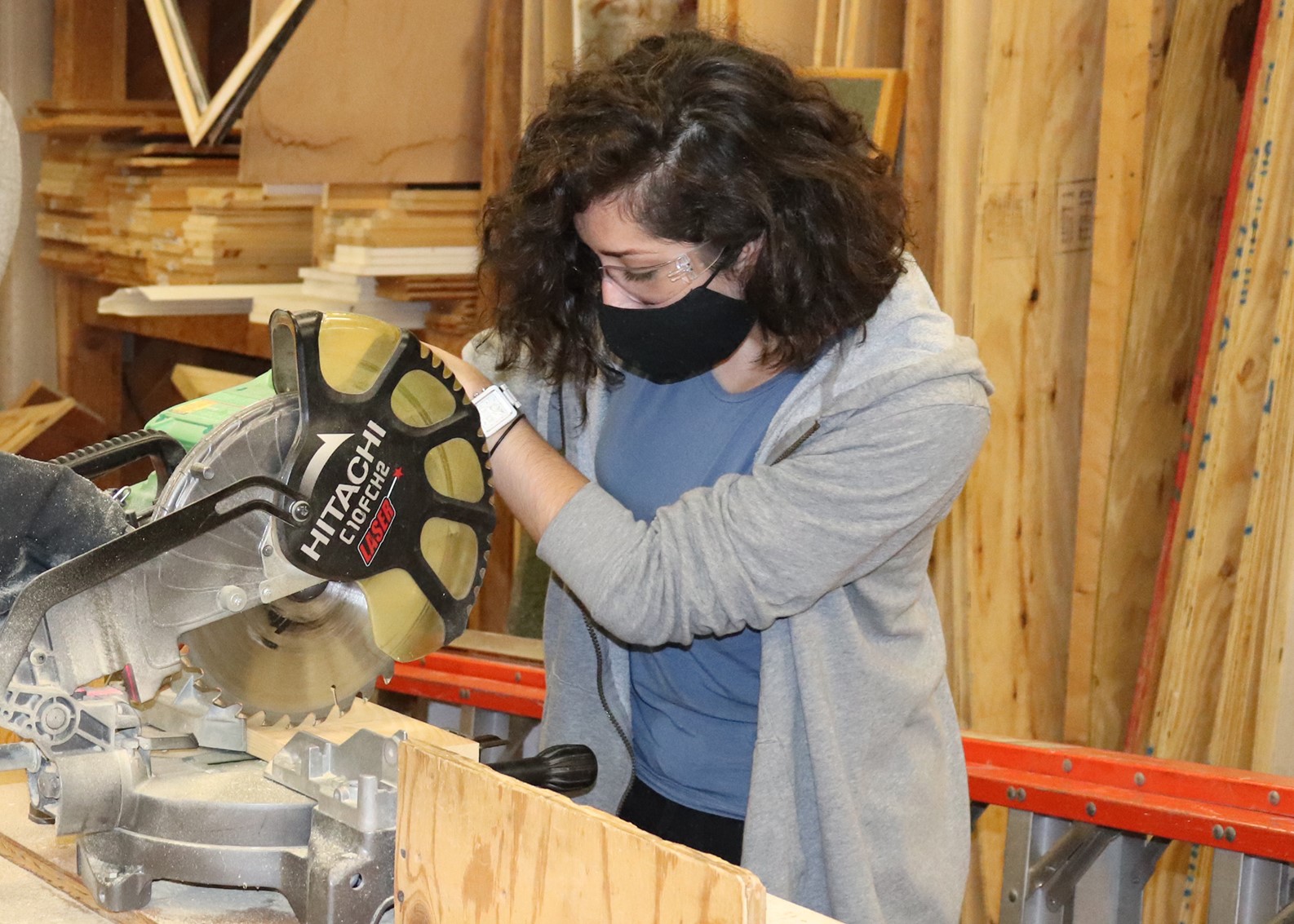 Student Isabel Tribunella. wearing protective eyewear and a face mask, uses a miter saw to cut through a board in a Capital Region BOCES Building Trades classroom.