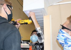 Three students wearing face coverings and protective eyegear use a drill to install a ceiling panel in a Capital Region BOCES Construction/Heavy Equipment classroom.