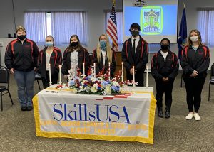 Five Albany Career and Technical School Skills USA officers, wearing their blue and red uniform, stand in a row behind a table draped in a SkillsUSA banner and holding flowers and candles.