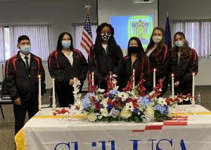 Six Albany Career and Technical School Skills USA officers, wearing their blue and red uniform, stand in a row behind a table draped in a SkillsUSA banner and holding flowers and candles.