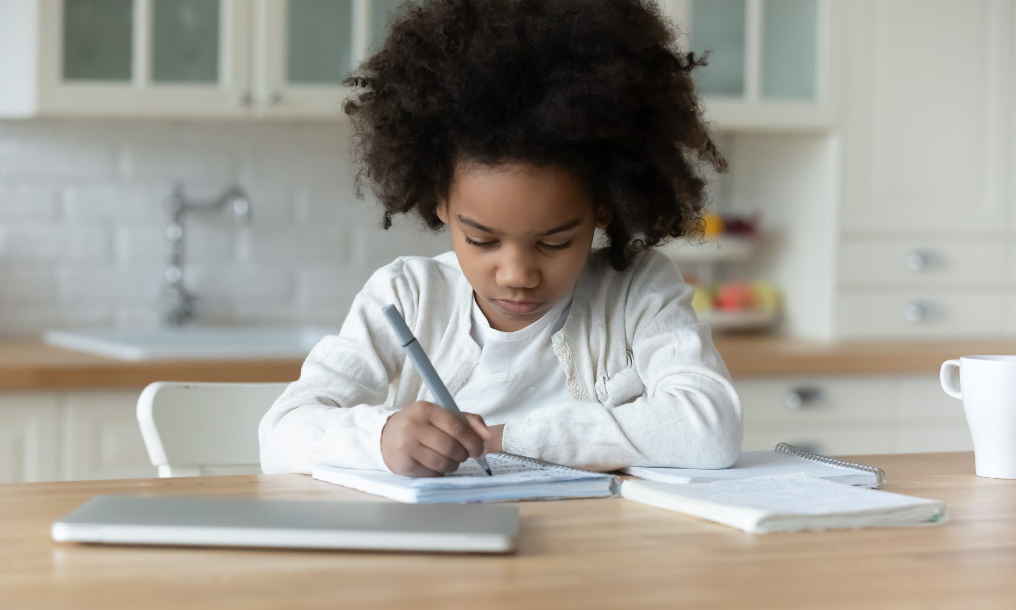 A young girl wearing a white t-shirt and cardigan sweater and holding a pen, sits at a kitchen table and writes in a spiral-bound notebook.