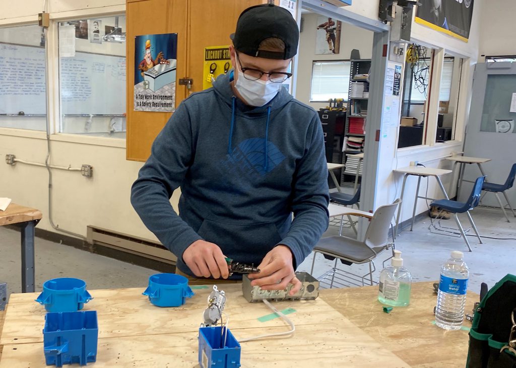 An adult student wearing a black ball cap turned backwards, glasses and a face mask works with wiring at a workbench in an Electrical Trades classroom. 