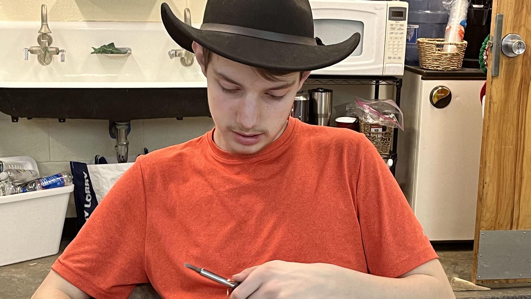 Adult Education students Griffin Boehlke, who is wearing a brown felt cowboy hat and a short-sleeved orange t-shirt, is sitting at a work table, and looking downward while bending wire with a hand tool.