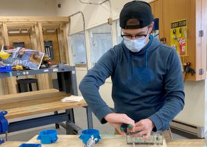 Adult student Kevin Felton, wearing a black ball cap turned backwards, glasses and a face mask works with wiring at a workbench in an Electrical Trades classroom.