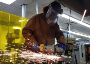 Adult Welding and Metal Fabrication student Laura Kessler, wearing a face mask and heat protective clothing and a shield welds metal as sparks fly in a Capital Region BOCES classroom. 