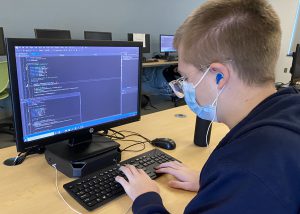 A seated student wearing a dark blue sweatshirt, eyeglasses and a light blue face mask works on laptop on a page of web code in a Career and Technical School Game Design and Implementation classroom.