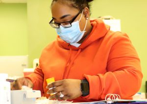 Adult Nursing student Sonisha Robinson, wearing eyeglasses, a light blue face mask and orange hooded sweatshirt, measures medicine in a Capital Region BOCES classroom.