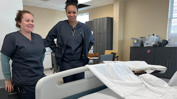 Two LPN students, Cassidy Knapp and Shenella Alert, wearing grey scrubs, look at a hospital bed.