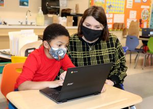 A Capital Region BOCES student wearing a bright red sweatshirt and protective face covering, and a teacher wearing a black and yellow plaid sweater and face covering, sit together at work on a Chromebook.