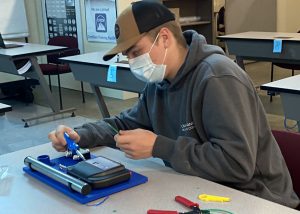 A student wearing a tans and black ball cap and protective face covering works on an electrical panel in a Capital Region BOCES Network Cabling classroom.