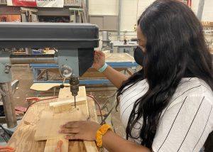 Capital Region BOCES student Enid Waring, wearing a white t-shirt with thin blue stripes and a protective face covering, works on a machine in a Building Trades classroom.