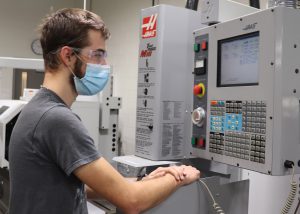 A student wearing a short sleeved grey t-shirt, protective glasses and a light blue protective face covering works on a computerized panel in a Capital Region BOCES Manufacturing and Machining Technology classroom.