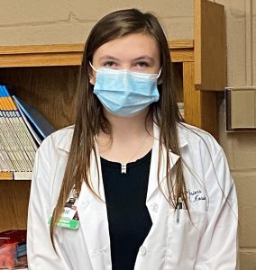 Capital Region BOCES New Visions: Health Careers student Karrie Kosier, wearing a long white medical coat and a blue protective face covering poses for the camera.