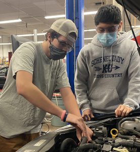 Two students  wearing grey shirts and protective face coverings work together over the engine of a car in a Capital Region BOCES Auto Trades classroom.