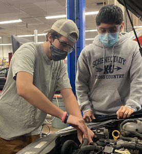 Two students wearing grey shirts and protective face coverings work together over the engine of a car in a Capital Region BOCES Auto Trades classroom.