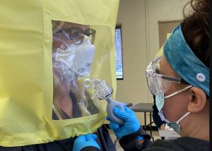 An adult nursing student wearing navy blue scrub top and bright blue hair wrap, tests the fit of a protective n95 face mask on another Adult Nursing student, wearing eyeglasses and a protective n95 face mask and surrounded by a bright yellow enclosure with a see-through window. 