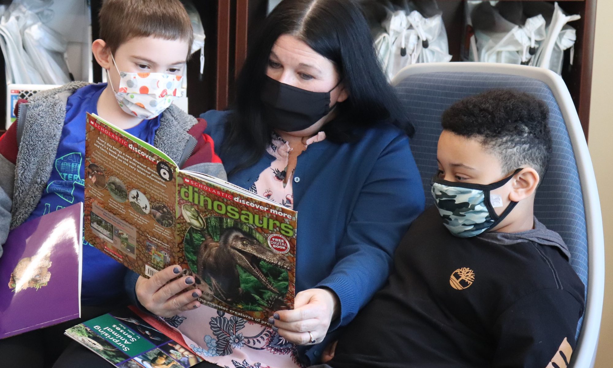 Two young students, one wearing a grey hoodie and face mask, the other wearing a black hoodie and face mask, sit on either side of a teachers with long black hair and a black face mask. The are reading a picture book on dinosaurs together.