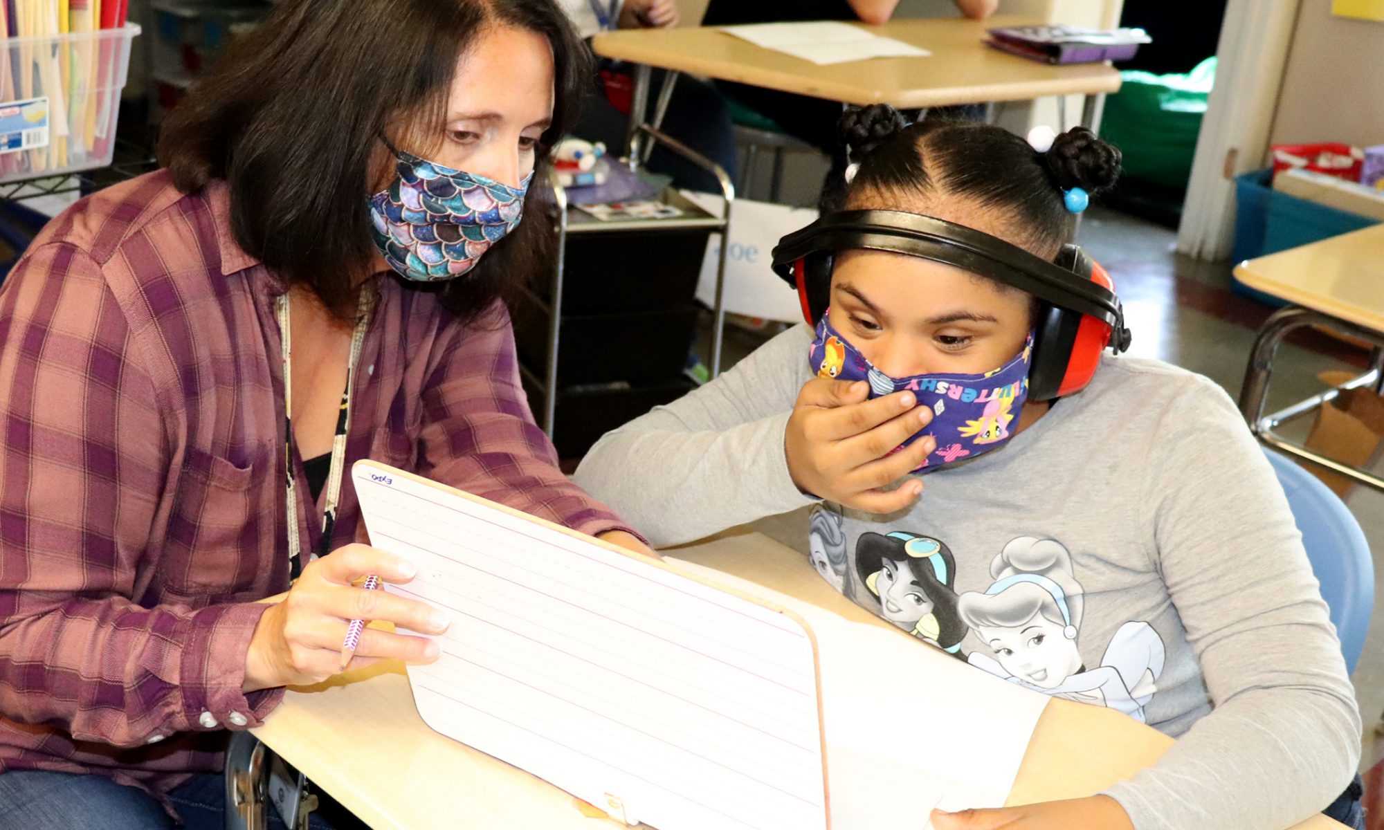 A teacher wearing a rust and orange plaid shirt and blue patterned protective face covering and a student wearing a sound muffling headset, protective purple patterned face covering and grey shirt work together on a Chromebook in a Capital Region BOCES special education classroom.