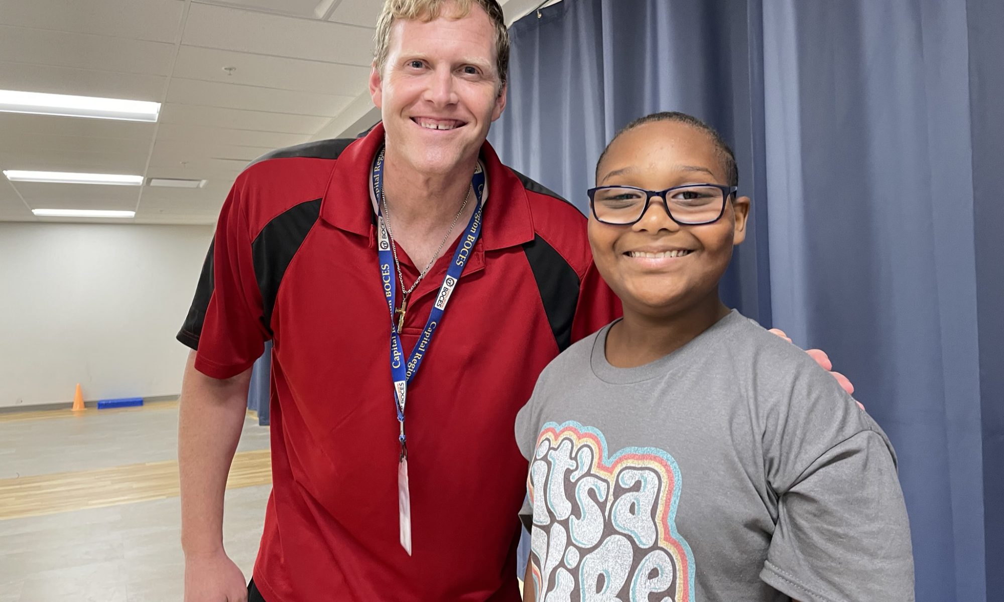 A staff member and student smiling. The staff member is wearing a red polo and the student is wearing a grey shirt that says "it's a vibe" in white letters.