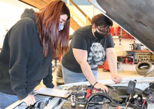 Auto Trades Technology students Makaya King, who has long auburn hair and is wearing a protective eyeglasses, an American flag printed face mask and a black hoodie, and Destiny Honsinger, with hair pulled back and wearing protective eye glasses, a black face mask and Marilyn Monroe skull t-shirt, inspect the engine of a car in a Capital Region BOCES auto lab.