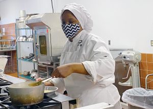 A student wearing a white chef's hat, apron and uniform, and a black and white zig-zag patterned protective face mask stands at a stove stirring food in a pot in a Capital Region BOCES culinary kitchen classroom. 