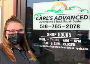 Capital Region BOCES Diesel Tech program alum Victoria Carl, who has long hair and is wearing eyeglasses, a black hoodie and black protective face covering, stands alongside the sign for Carl's Advanced Automotive and Truck Repair Center and smiles for the camera.