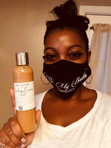 Capital Region BOCES Cosmetology alumna Tiffany Harris, wearing a black protective face mask embroidered with her company name, Whip My Butta, and a white v-neck t-shirt, holds a bottle of skin care and smiles for the camera.