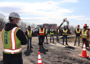 Capital Region BOCES Construction and Heavy Equipment students stand outdoors under a blue sky and in the dirt in a semi-circle facing a representative from BBL Construction, the firm that is constructing the new Career and Technical School Albany campus and Maywood School. All are wearing bright orange hard hats, bright yellow reflective vests and protective face masks.