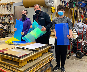 Two Capital Region BOCES Digital Media students, one wearing a purple hoodie and black protective face mask, the other with curly brown hair, a grey t-shirt and bright blue face mask, stand on either side of work-based learning coordinator Jim Haas. All three hold bright blue and neon yellow acrylic sheets, which were recently donated to the Career and Technical School.